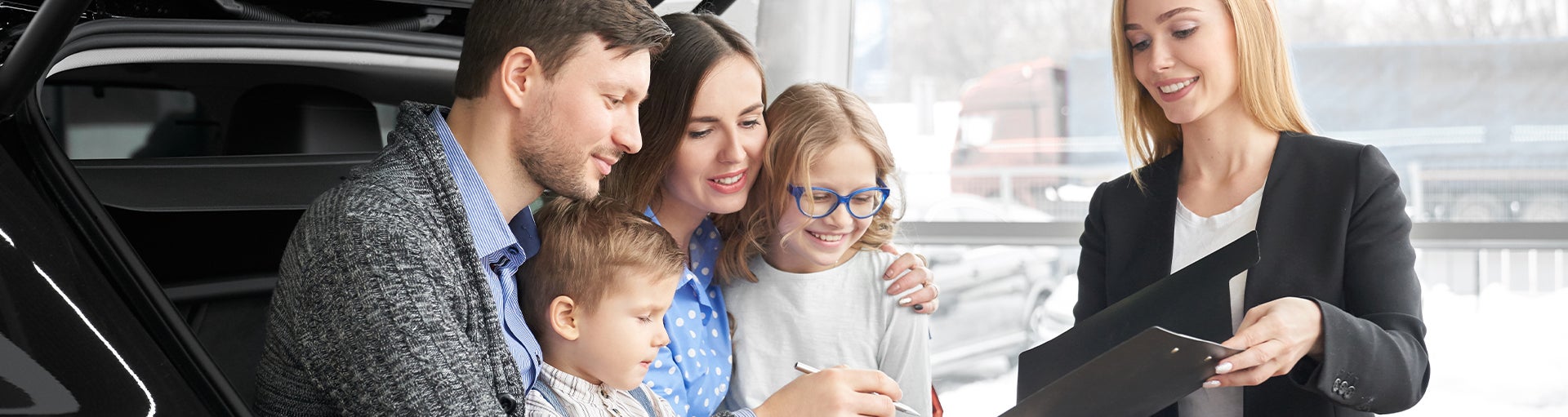 A family sitting in the back of a hatchback vehicle signing a document with a salesperson.