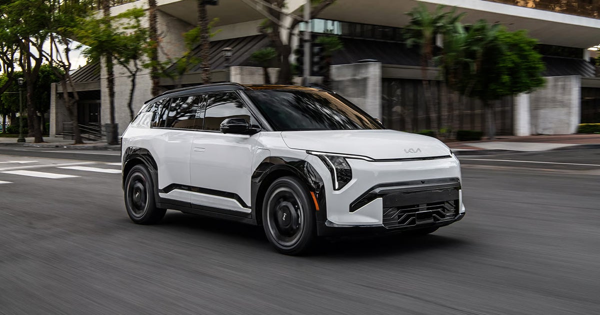A white 2027 Kia EV3 compact electric SUV driving on a city street.